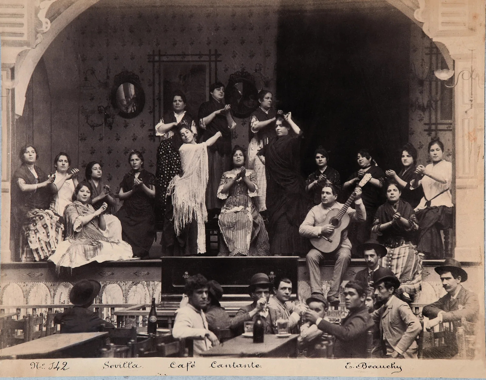A sepia print of a Seville café cantante around 1888, with women in shawls and polka-dot dresses seated and standing on a stage behind men at café tables