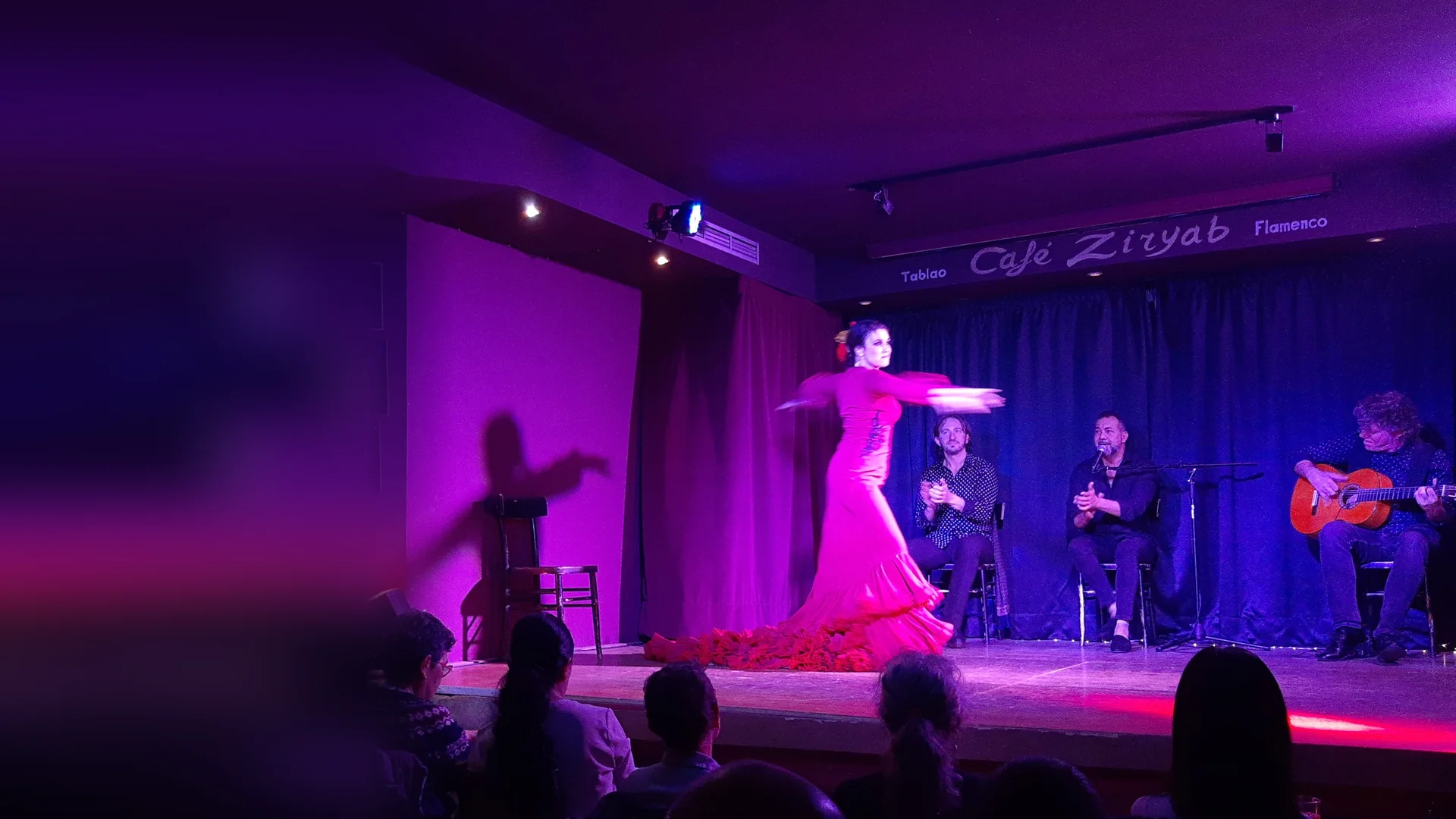 A dancer in a fuchsia dress mid-turn on the small stage of Café Ziryab in Madrid, with two seated artists clapping and a guitarist at the right