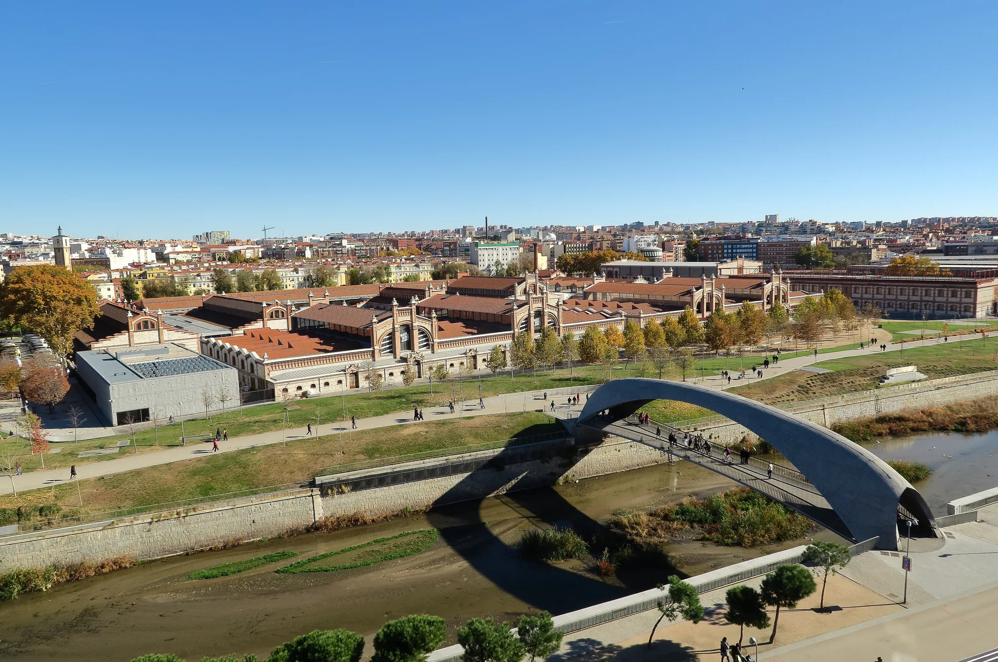 The Matadero Madrid buildings beside the Manzanares in Arganzuela, seen from the Madrid Río park under a clear blue sky, with a curved footbridge in the foreground