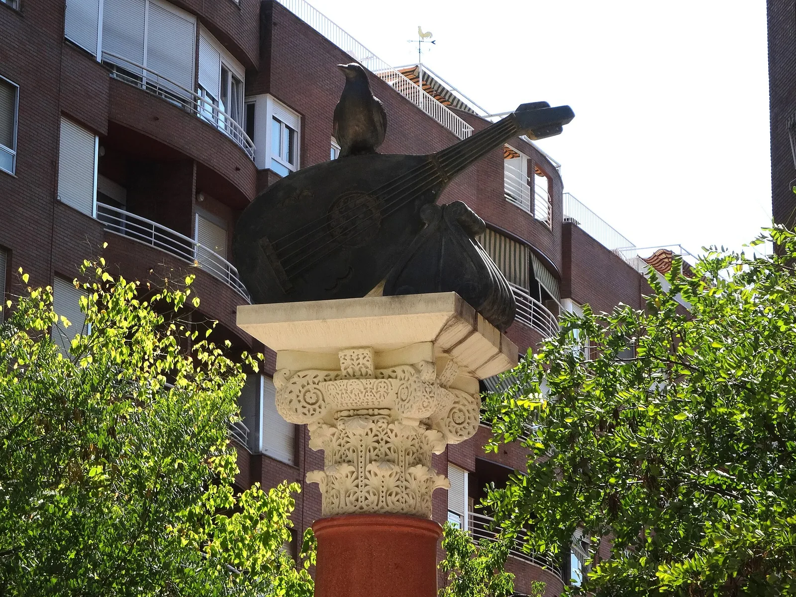 A dark bronze blackbird perched on a lute above a carved stone capital, the monument to Ziryab in Córdoba, with trees and apartment blocks behind