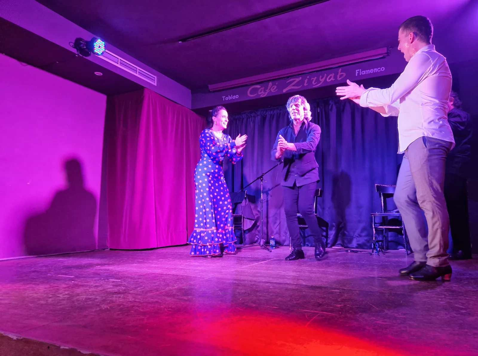 Three artists on the small stage of Café Ziryab under magenta light: a woman in a blue polka-dot dress and a man in a dark shirt clapping, and a man in a white shirt with raised hands